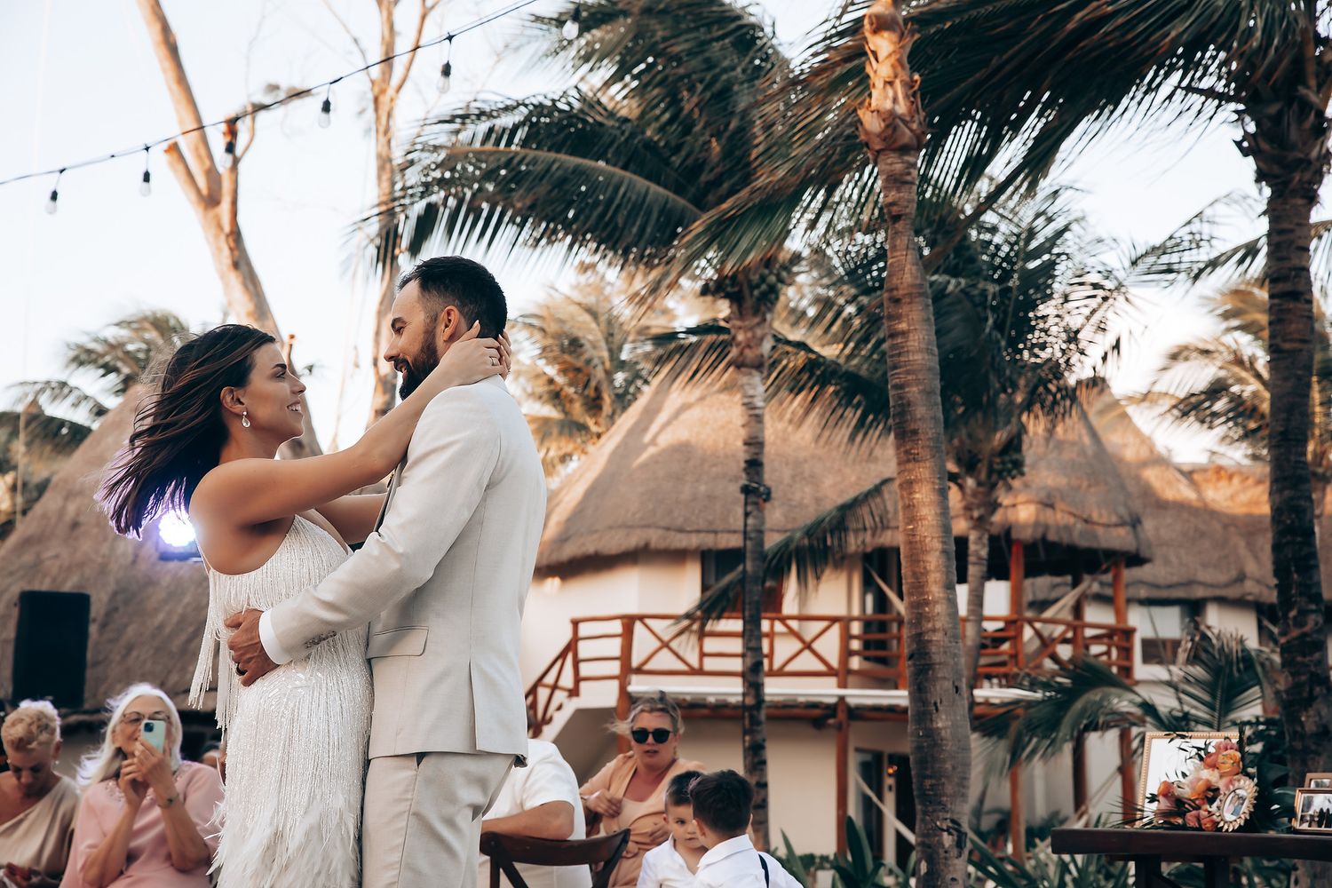 Wedding celebration under string lights with guests dancing in white attire among palm trees.