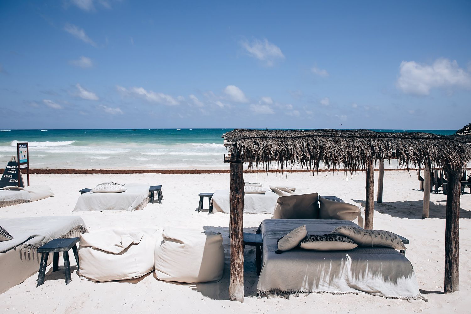 Luxurious beach bed with thatched canopy and beige cushions on white sand overlooking turquoise Caribbean waters.