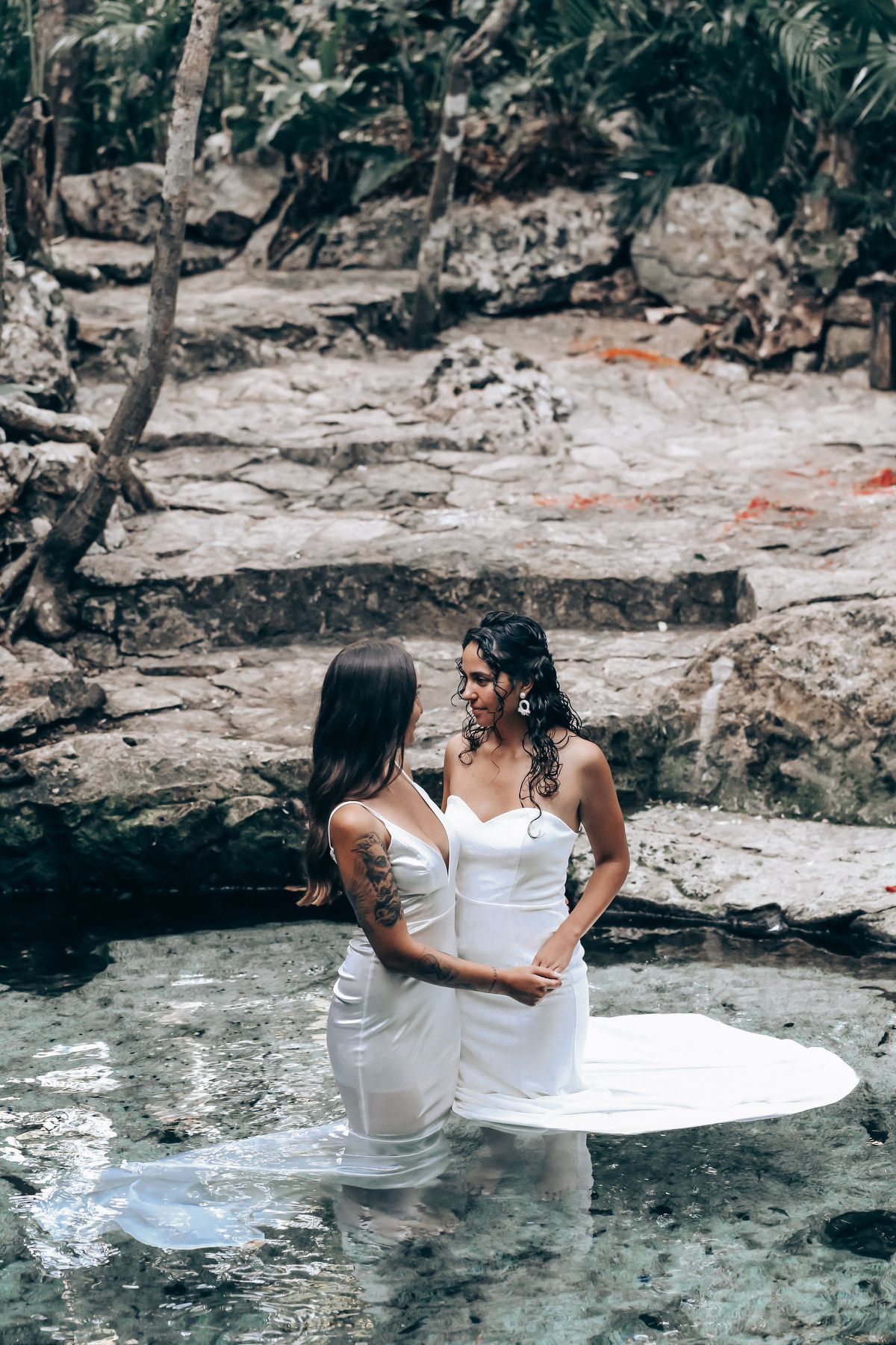 Two people in white dresses wade in crystal clear water surrounded by tropical scenery.