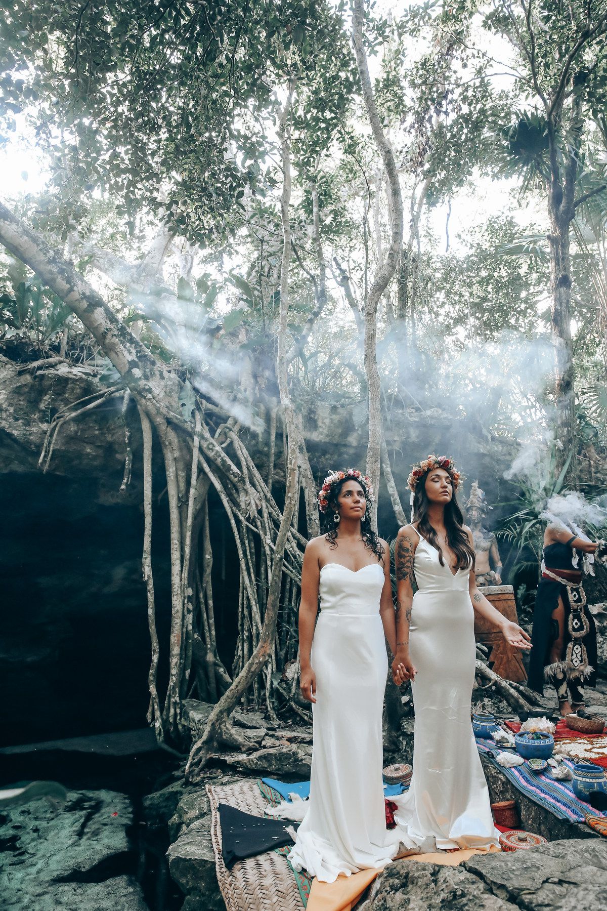 Two people in white dresses stand together in a mystical forest clearing with smoke effects.