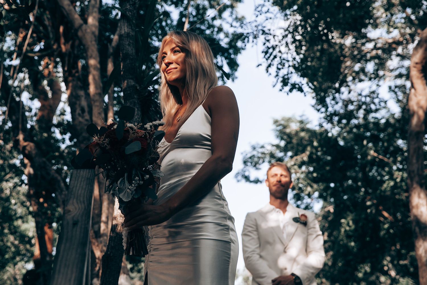 Wedding party in white attire stands together in a lush outdoor forest setting.