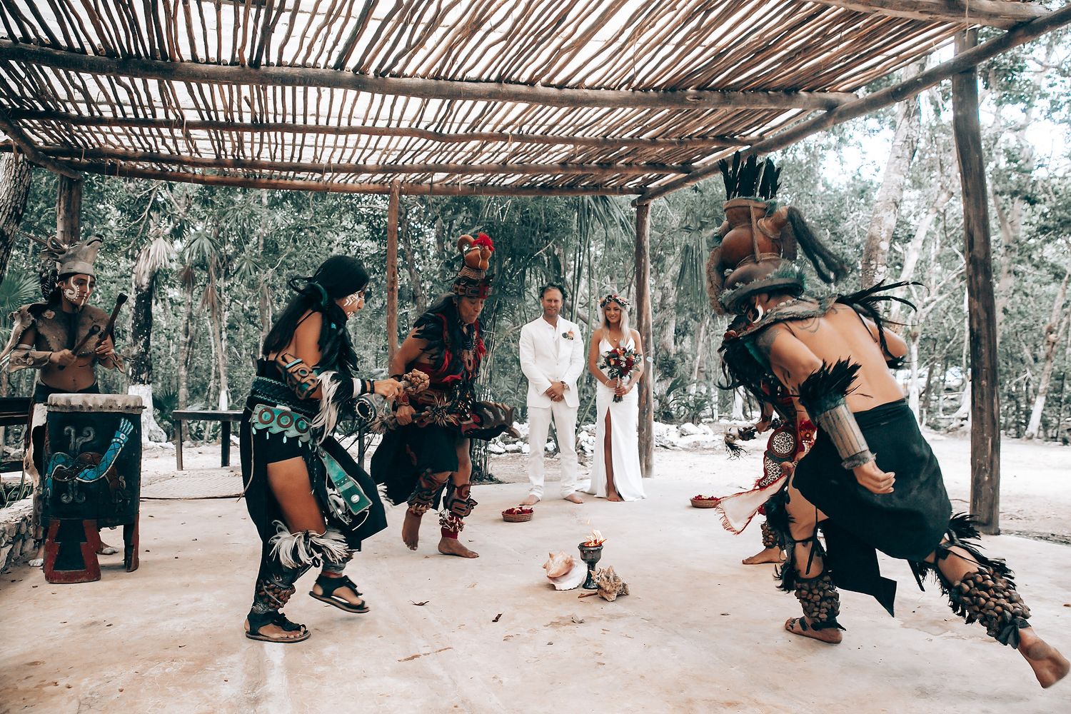 Traditional Mayan dancers performing ritual dance in ceremonial costumes under thatched roof.