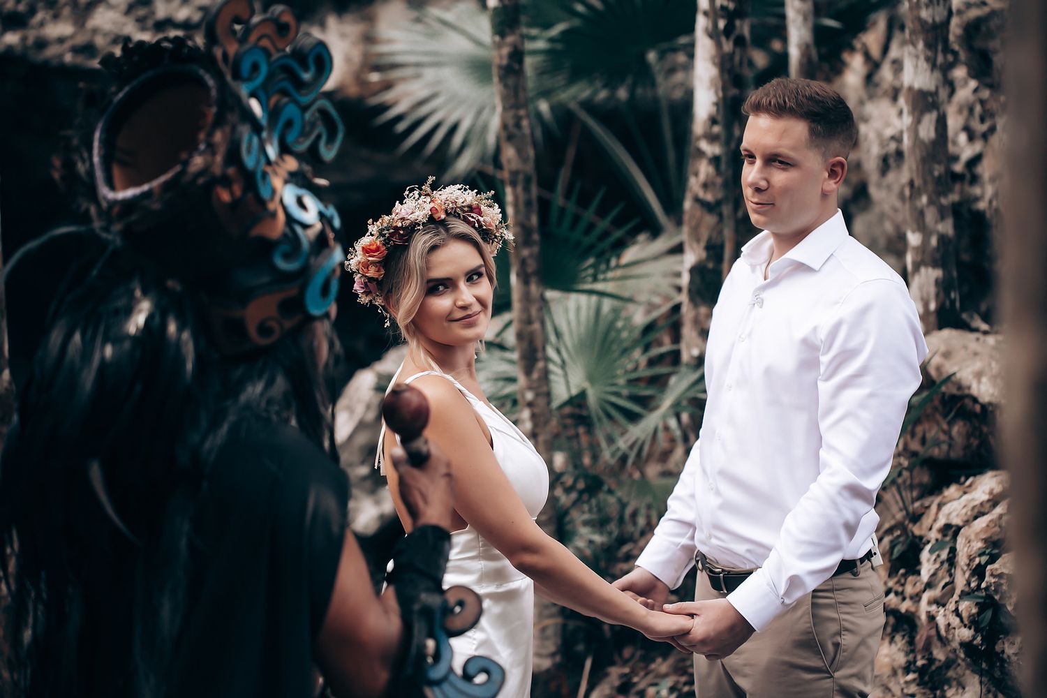 Couple in white attire sharing a joyful dance moment in a tropical garden setting with artistic wall decor.