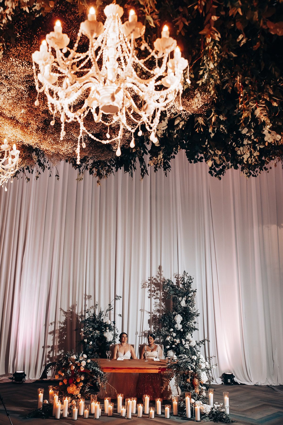 Elegant wedding reception setup with crystal chandeliers and floral arrangements against white draping.