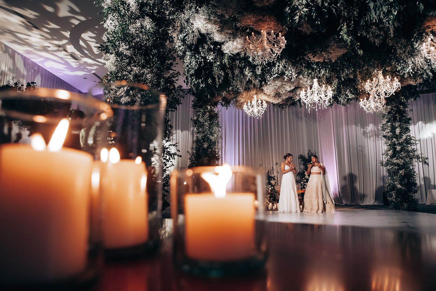 Romantic candlelit wedding reception decor with chandeliers and greenery overhead.