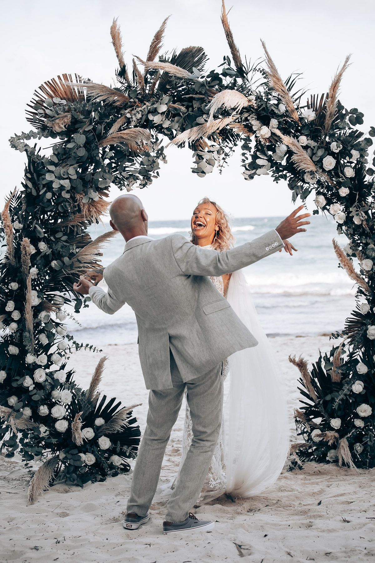 Newlyweds celebrating joyfully under a floral arch on a sandy beach.