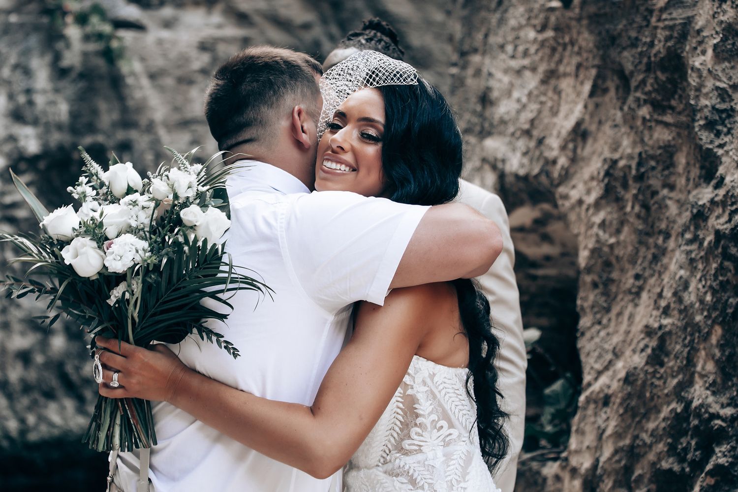 Newlyweds share a tender embrace while holding a white floral bouquet.