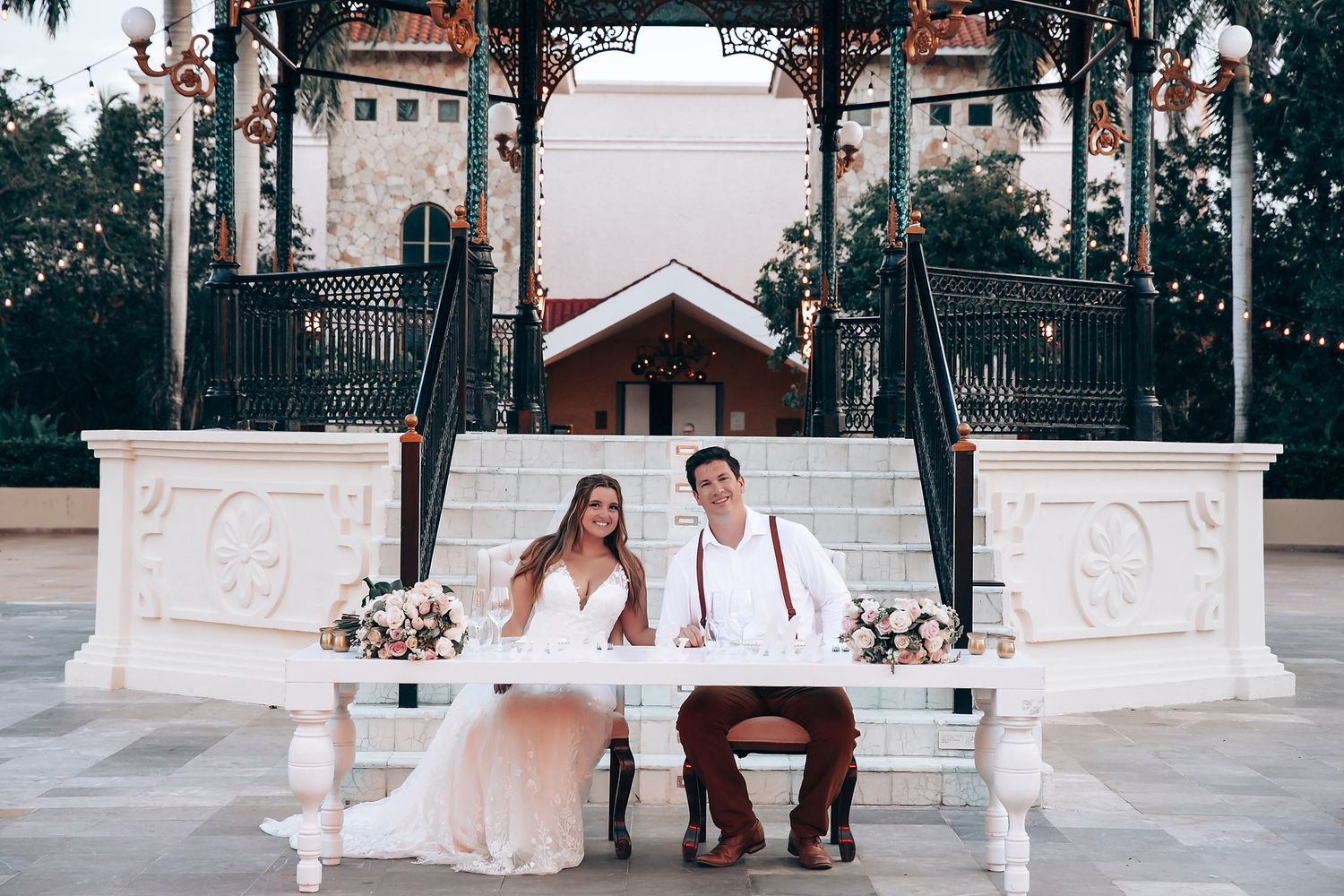 A couple sits on an ornate white bench at an elegant outdoor wedding venue with floral arrangements.