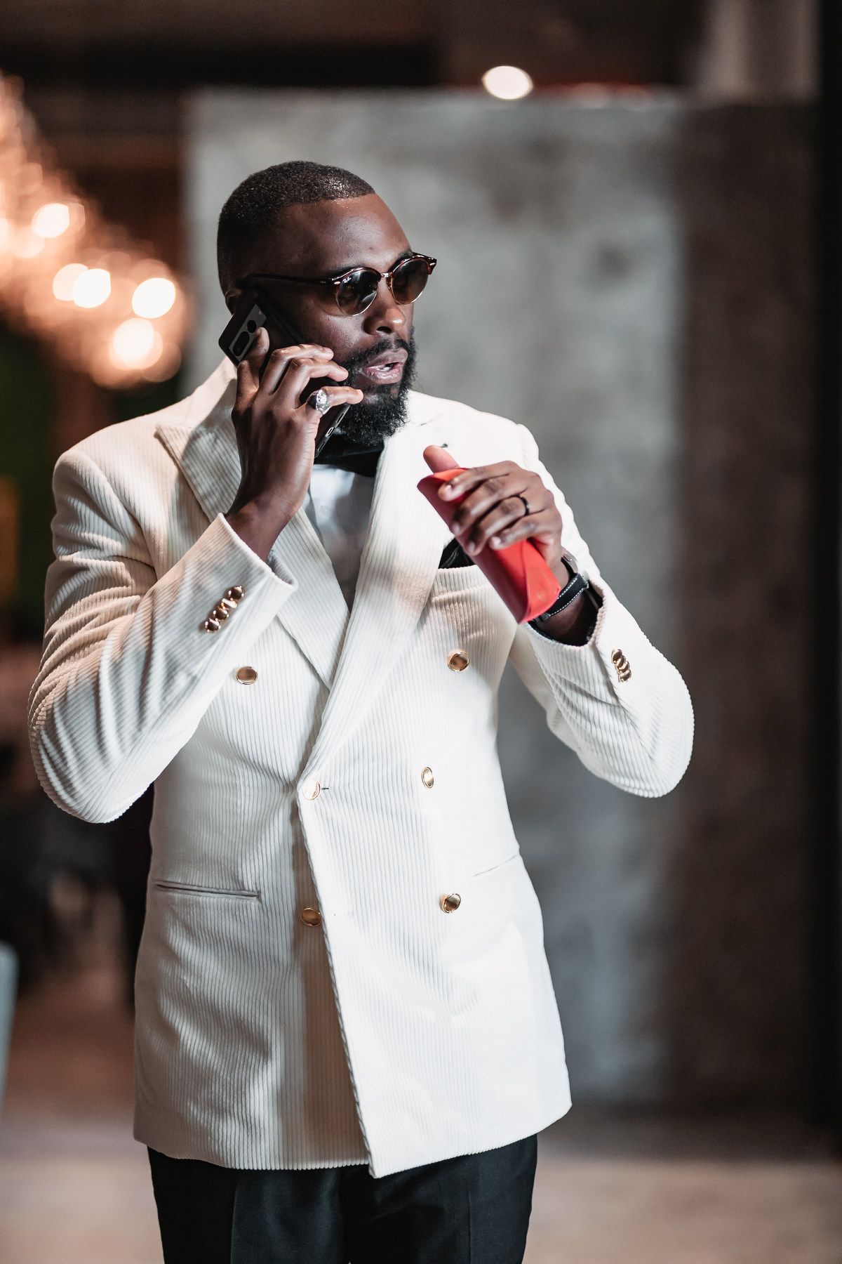 Stylish Black man in white double-breasted blazer and sunglasses talking on phone at elegant venue.