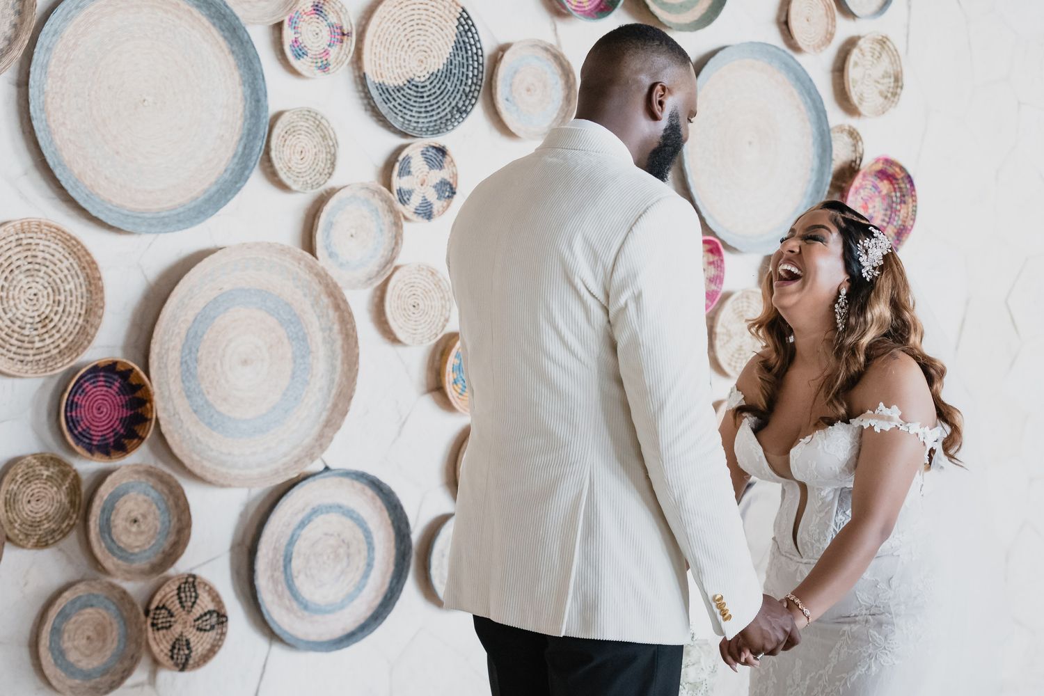 Bride and groom holding hands and laughing in front of a decorative woven basket wall display.