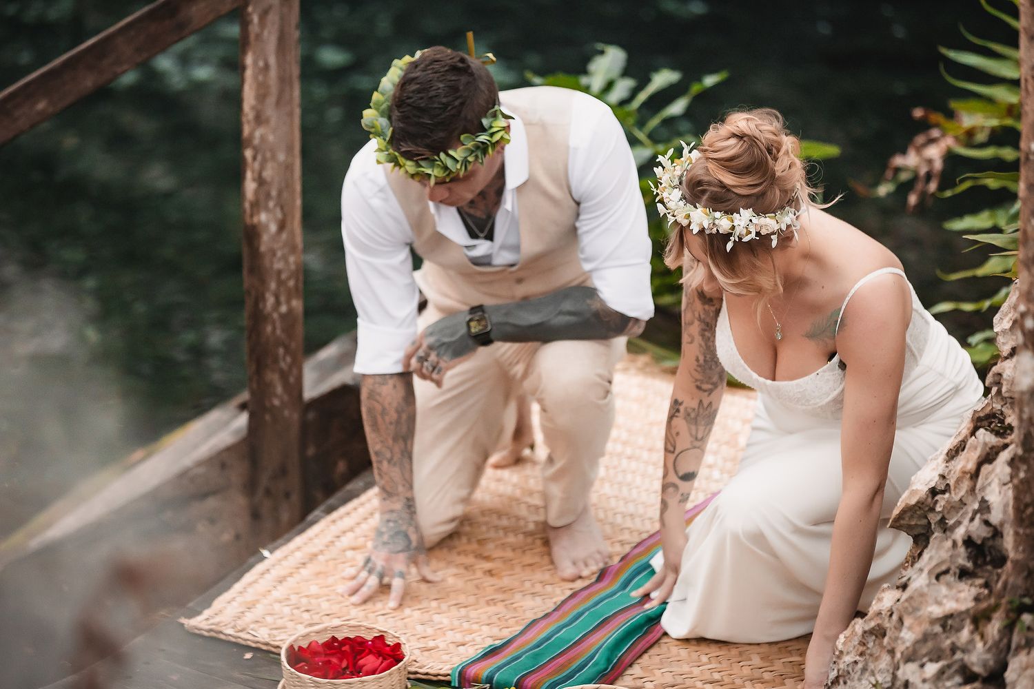 Couple in floral crowns performing a traditional wedding ceremony on a woven mat outdoors near lush greenery.