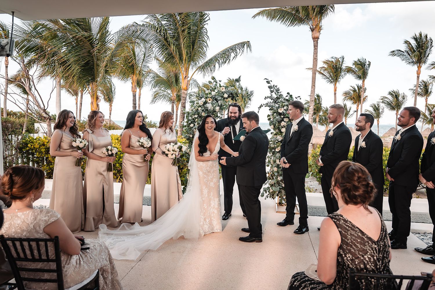Elegant outdoor wedding ceremony with bridesmaids in champagne gowns and groomsmen beside couple under palm trees.