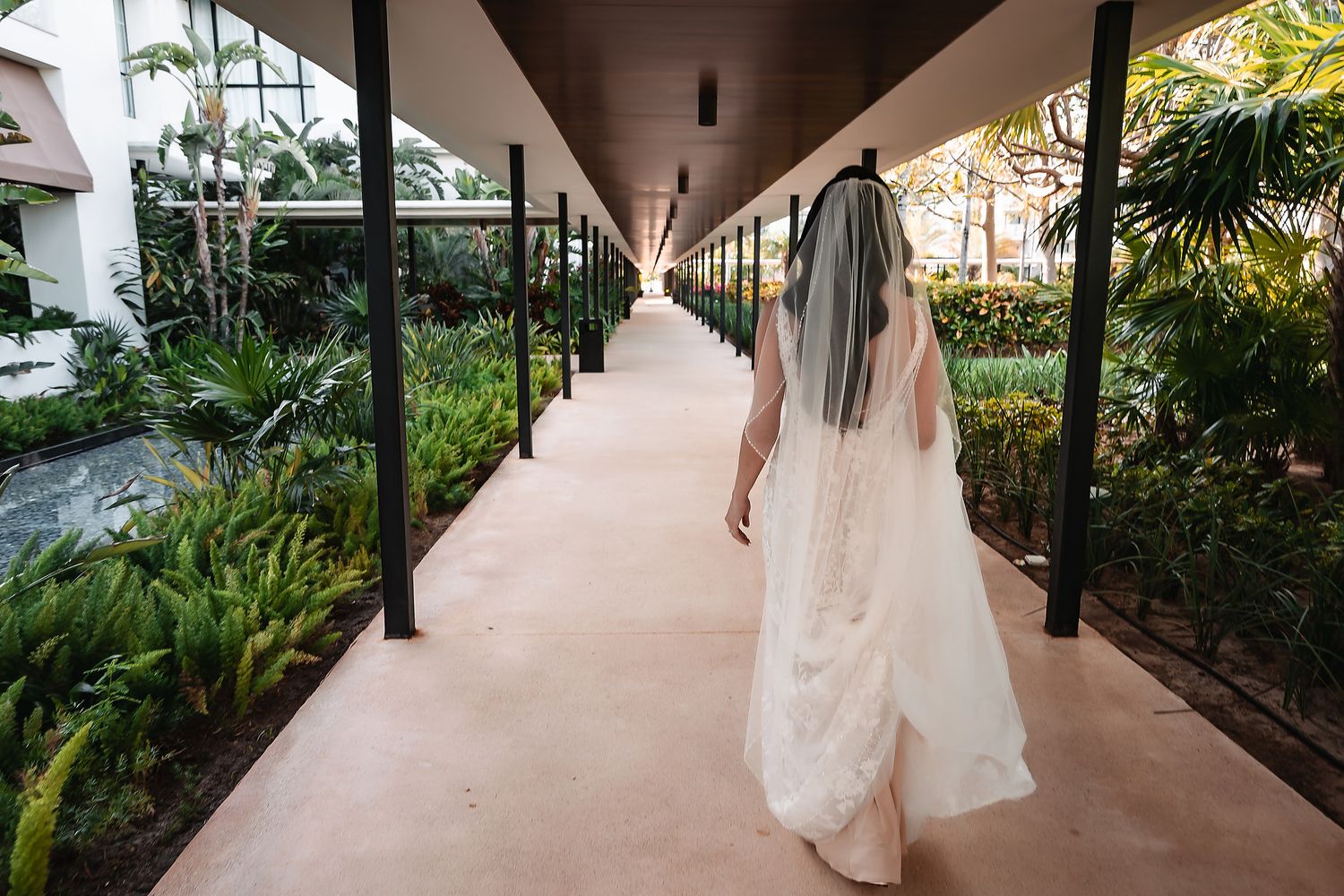 Bride in white wedding gown and veil walking down a lush tropical corridor at an elegant resort venue.