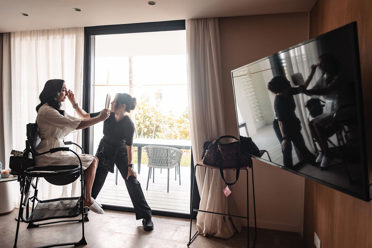 A makeup artist applies makeup to a client seated in a chair near a large window in a modern hotel room.