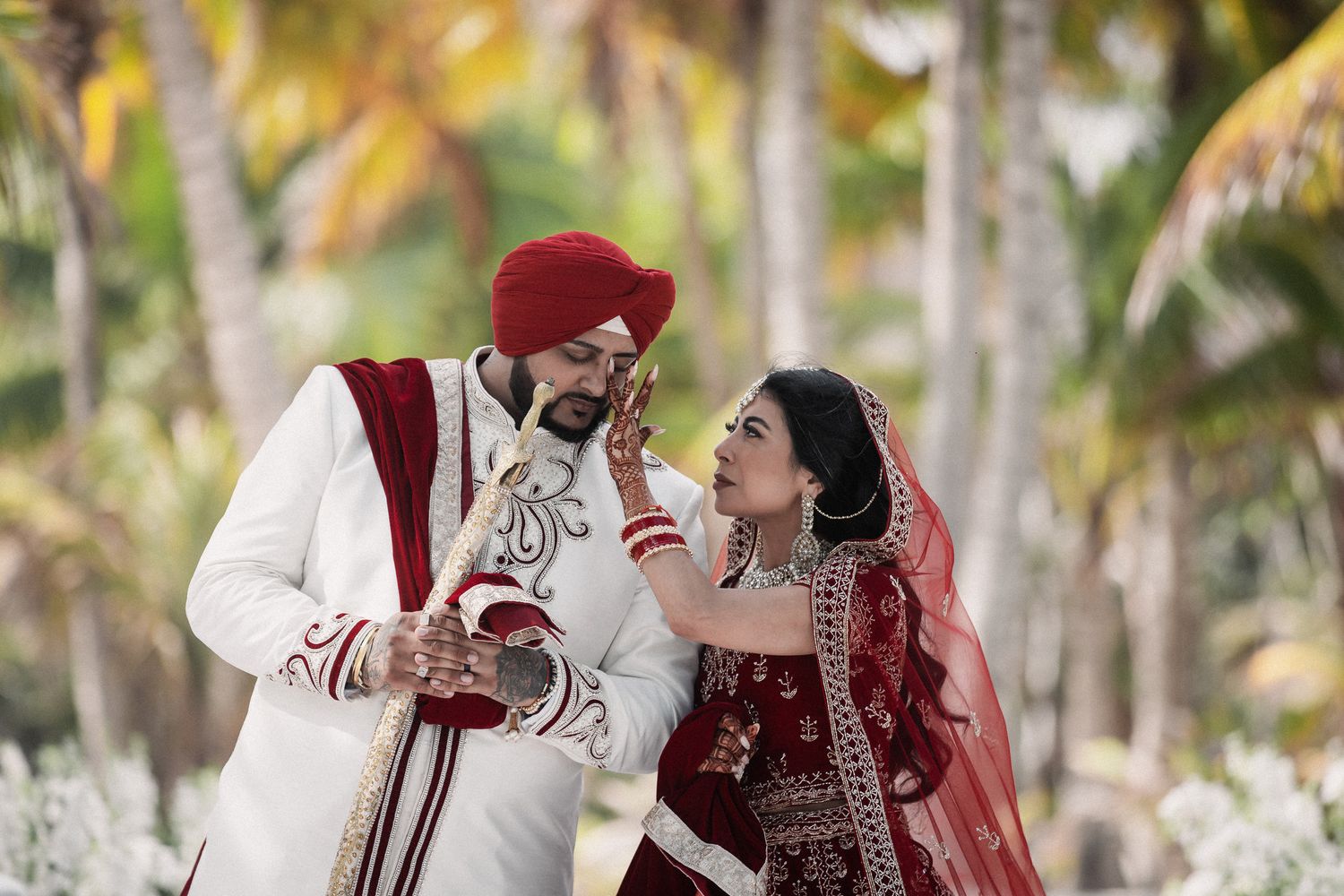 Indian bride in red bridal lehenga and groom in white sherwani sharing a tender moment surrounded by tropical palm trees.
