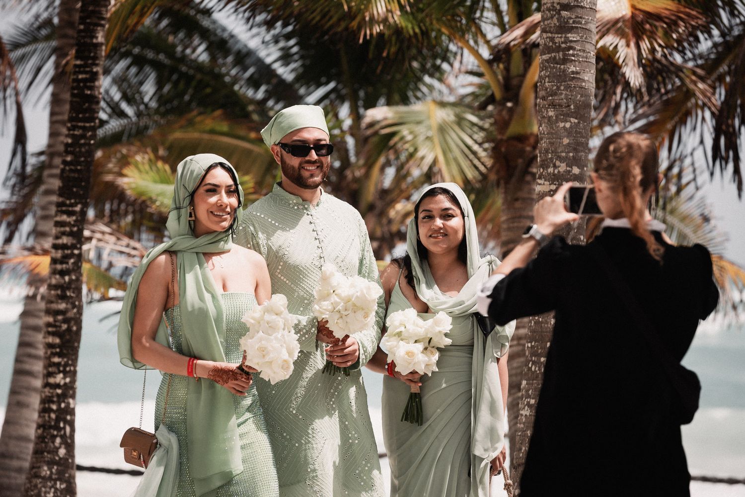 Muslim wedding party in mint green attire poses with white bouquets on a tropical beach.