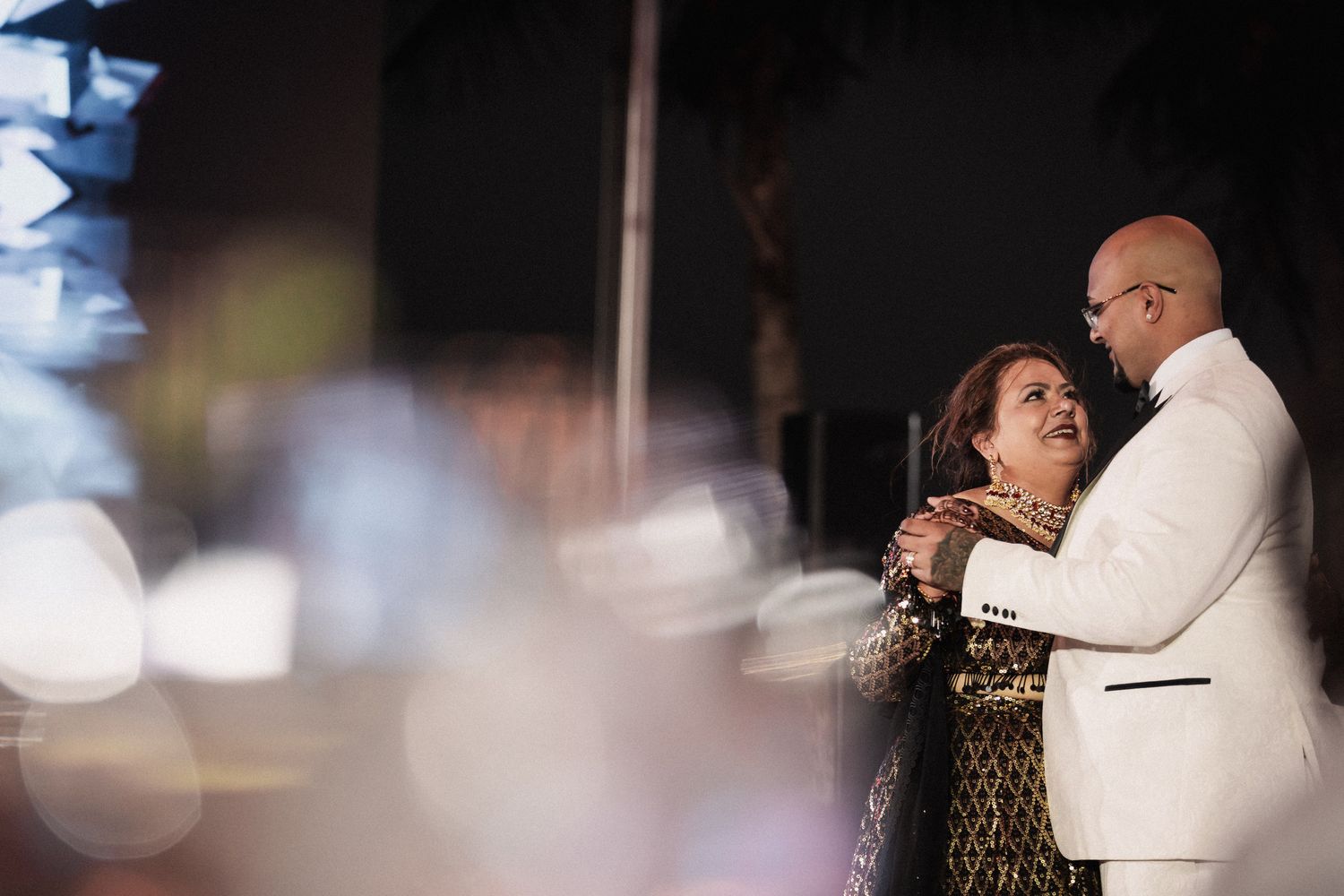 Groom in white tuxedo shares emotional mother-son dance at outdoor nighttime wedding reception.