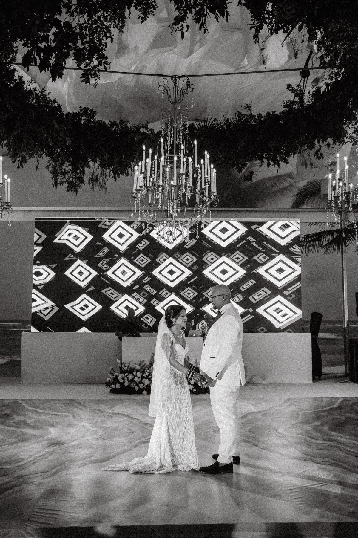 Black and white photo of bride and groom sharing first dance under chandelier at wedding reception.