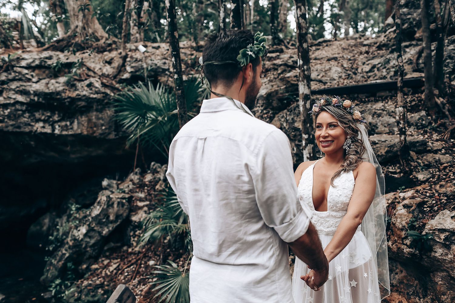 A romantic wedding ceremony takes place in a natural cenote cave setting with palm fronds in the background.