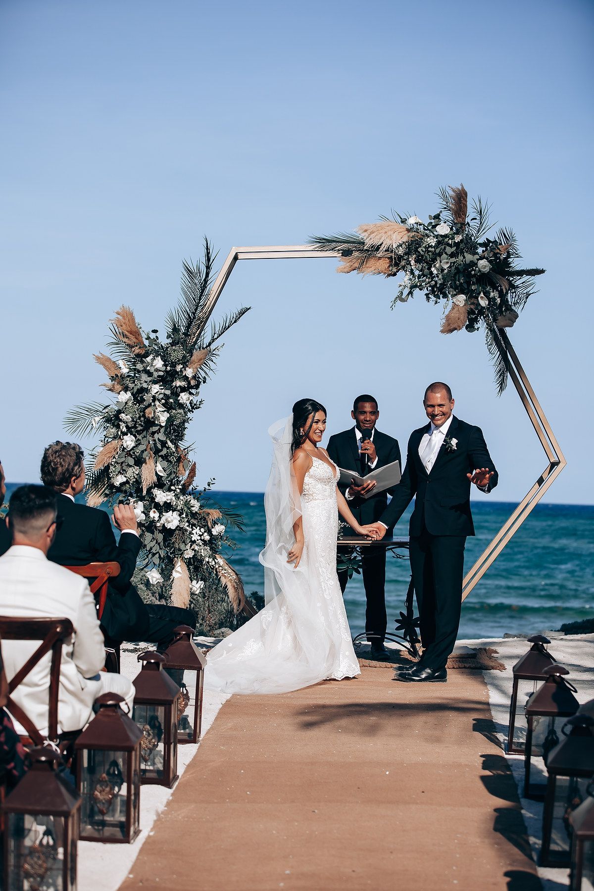 Beachfront wedding ceremony setup with wooden arch and floral decorations against blue ocean backdrop.