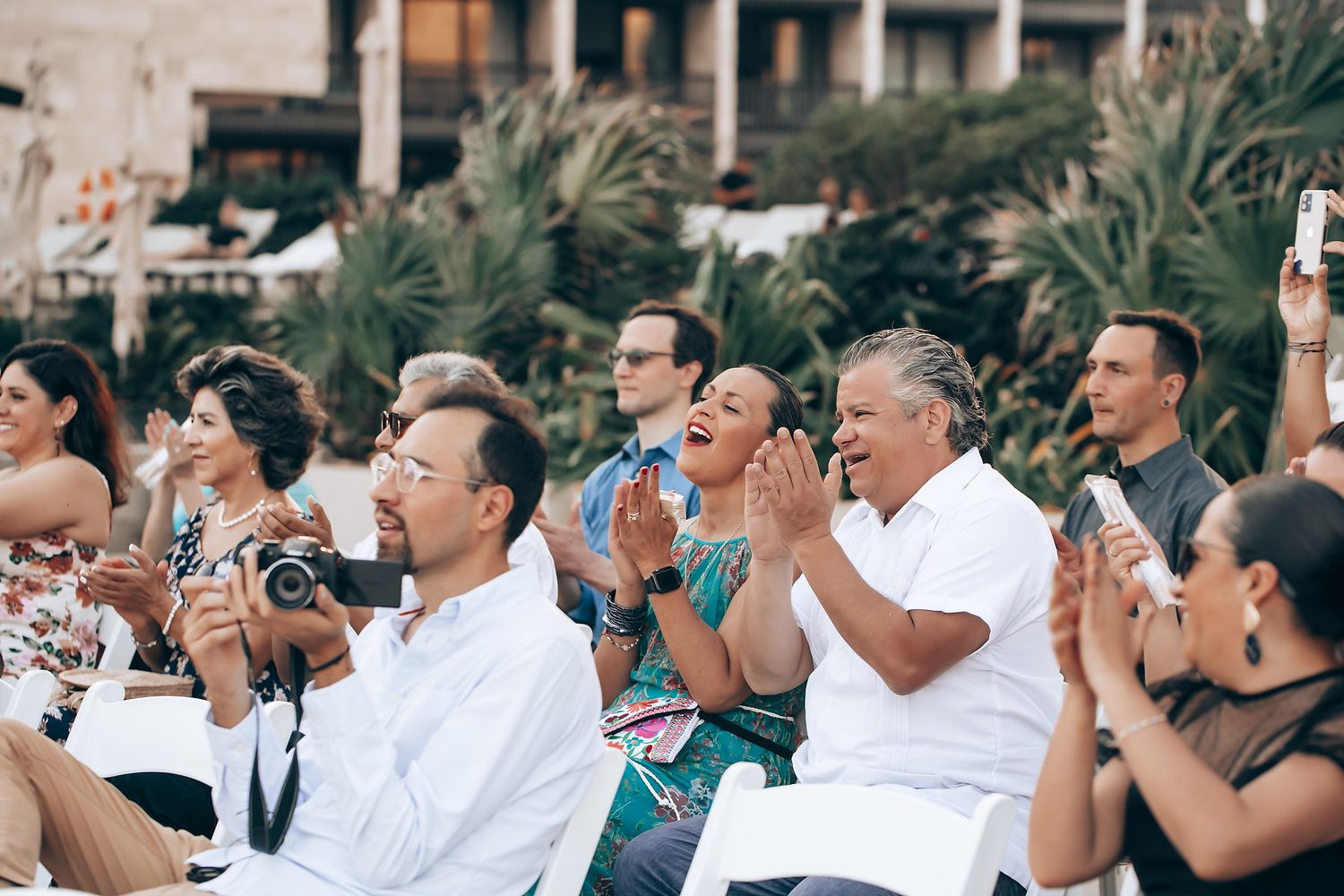 Audience members applaud during an outdoor event with palm trees in the background.