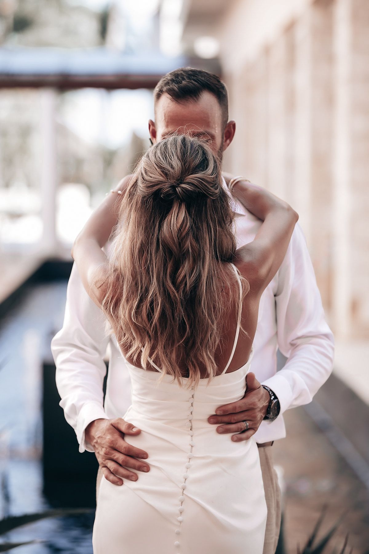 A woman with long, wavy hair is embracing a man from behind in a romantic setting.