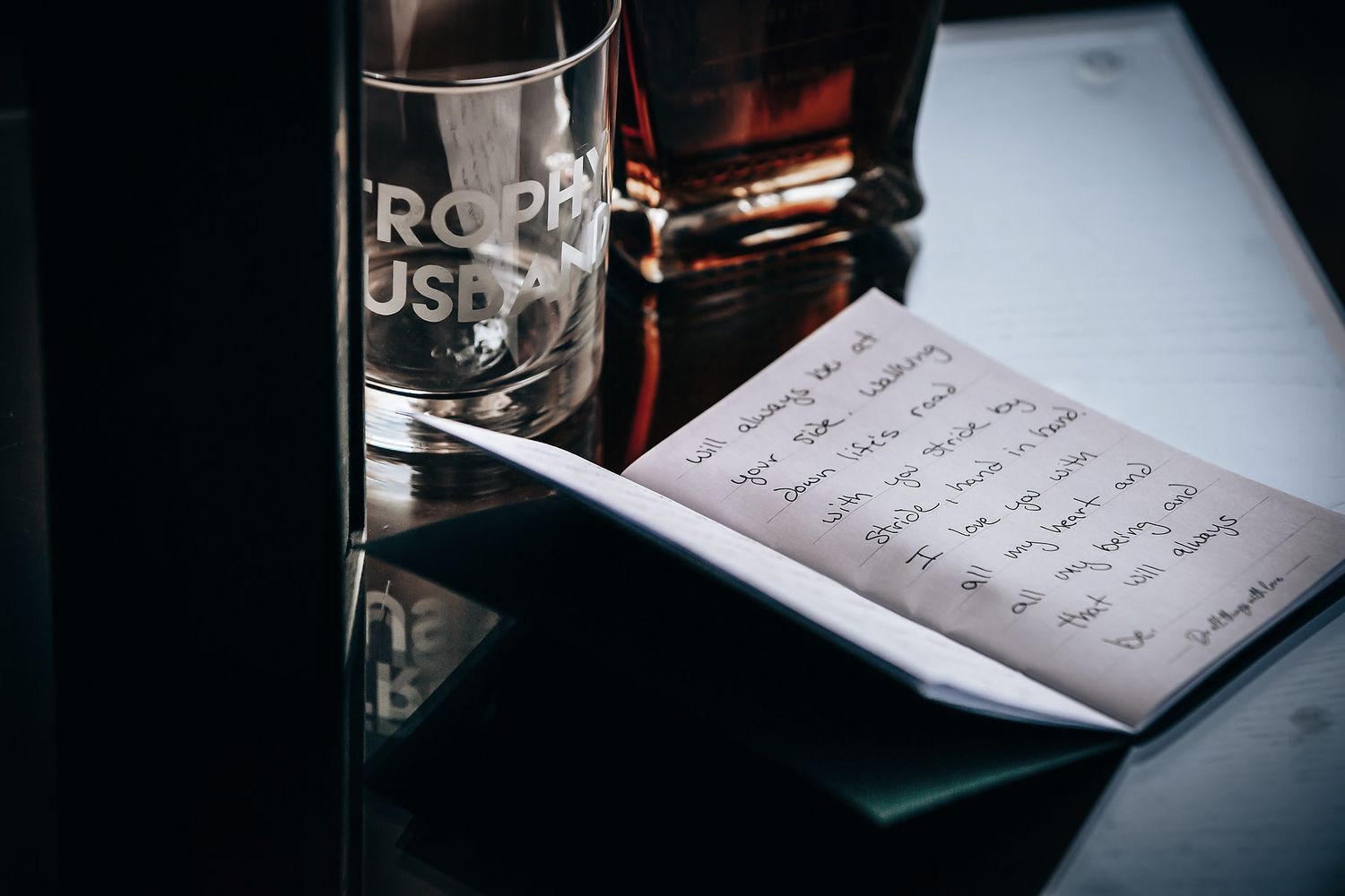 A glass of dark liquor and a notebook with handwritten text are placed on a reflective surface.