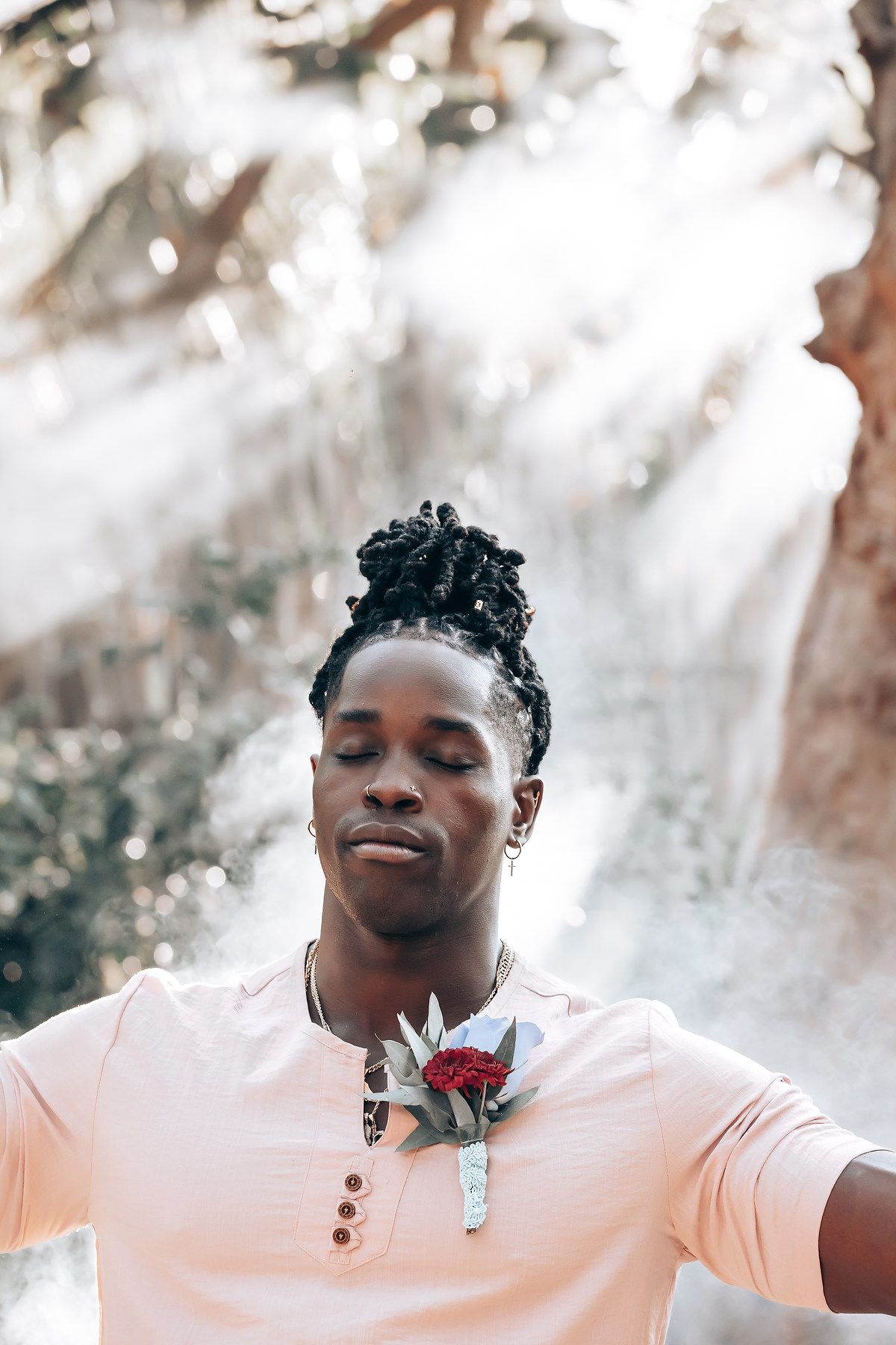 The image shows a smiling man wearing a pink shirt and black pants standing in a lush, green environment.