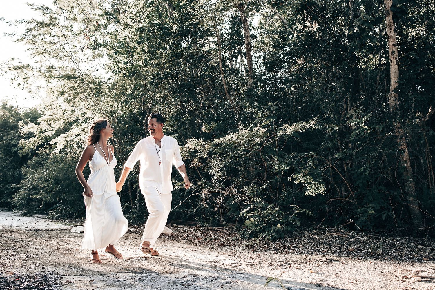 A couple walks hand-in-hand on a dirt path in a lush, green forest.