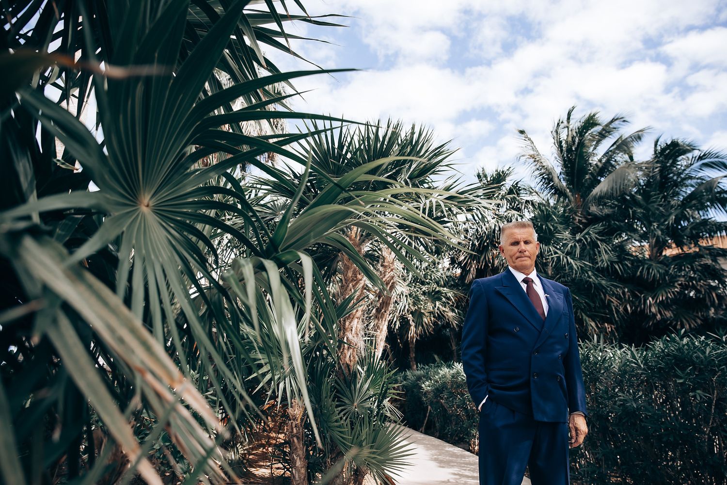 A man in a suit stands in a tropical setting with palm trees.