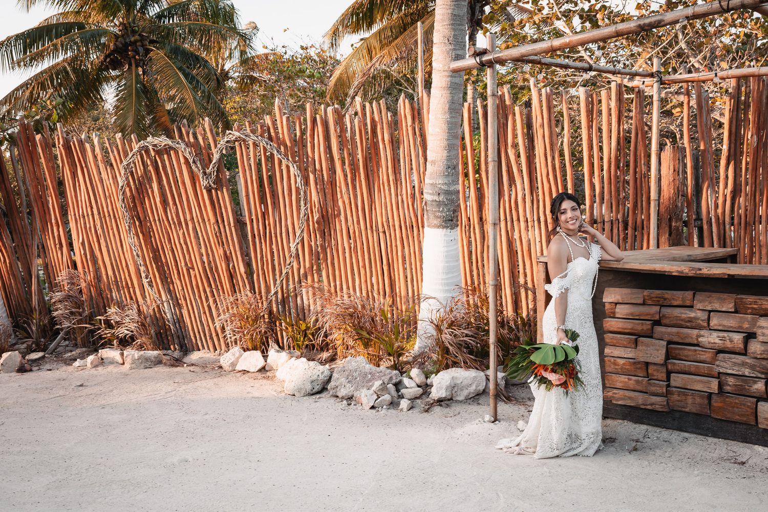 Bride in lace wedding gown holding bouquet beside rustic wooden arch on tropical beach at sunset.