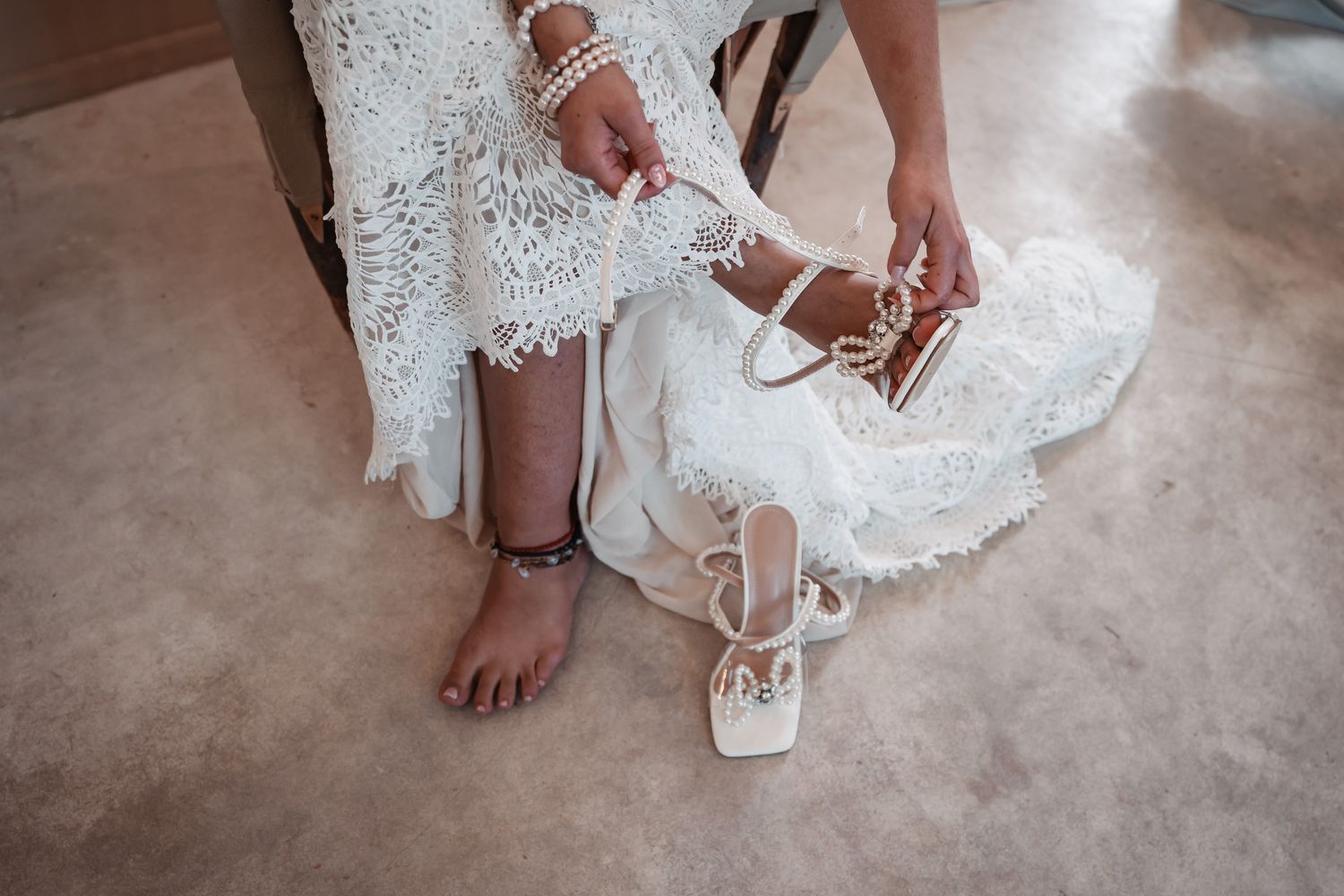 Bride in lace wedding dress putting on white strappy heeled sandals.