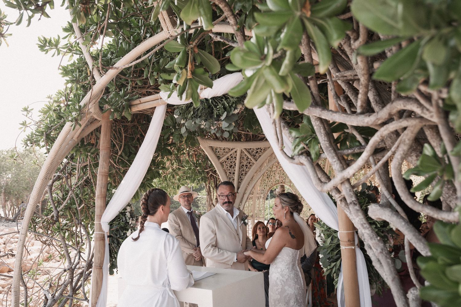 Bride and groom exchange vows under rustic driftwood arch adorned with white fabric outdoors.