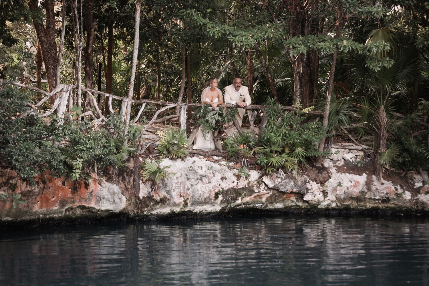 Newlyweds sit together on rocky cliff edge overlooking serene turquoise cenote in jungle setting.