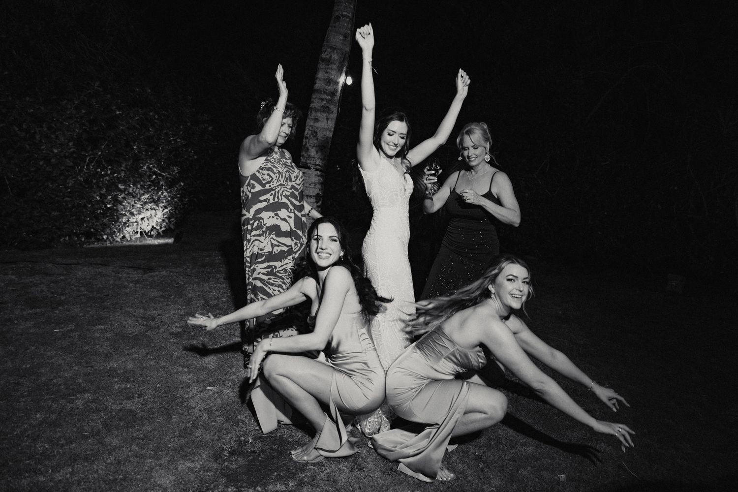 Bride poses with bridesmaids in playful black and white photo at outdoor wedding.
