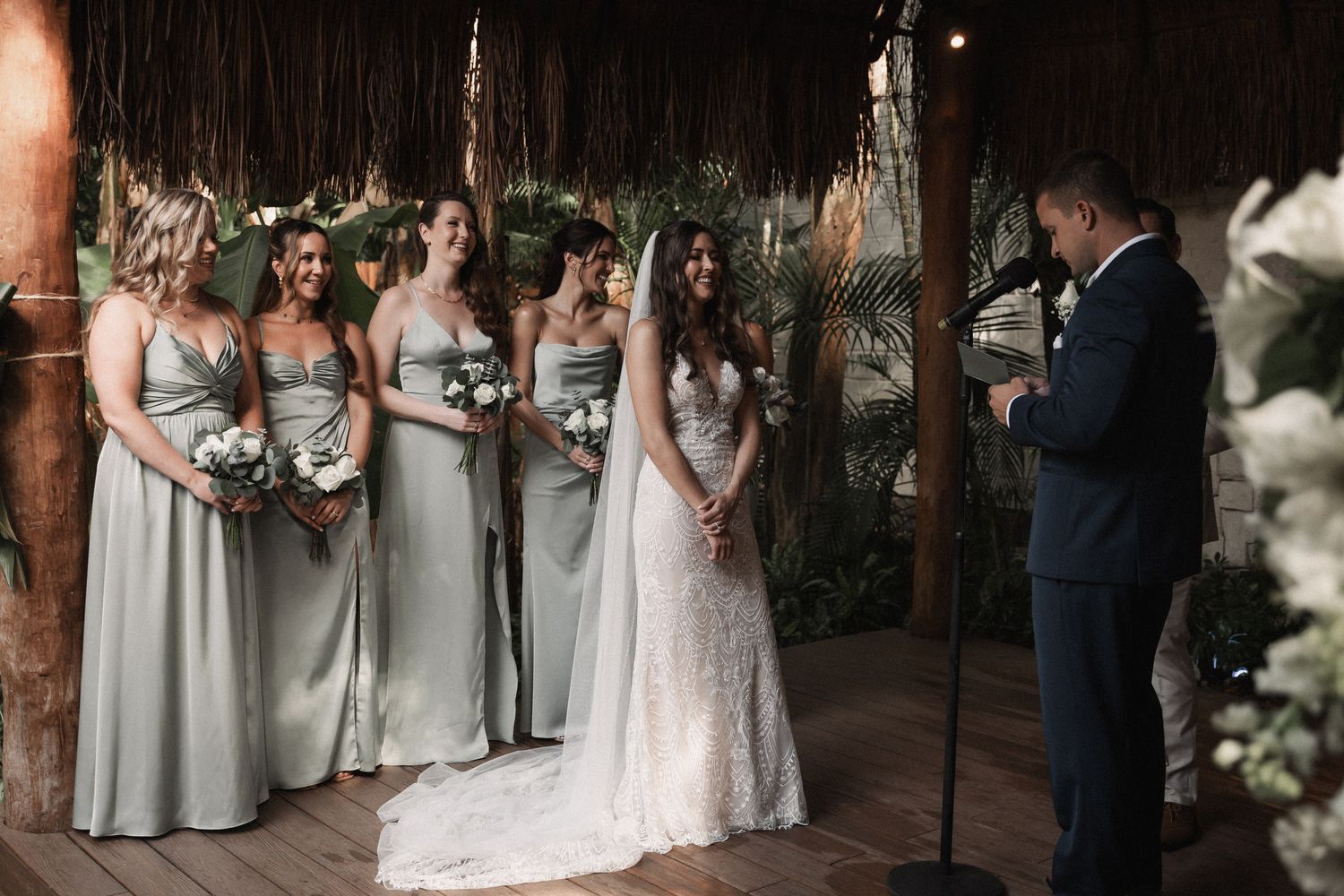 Bride in lace gown stands with bridesmaids in sage dresses during tropical wedding ceremony.