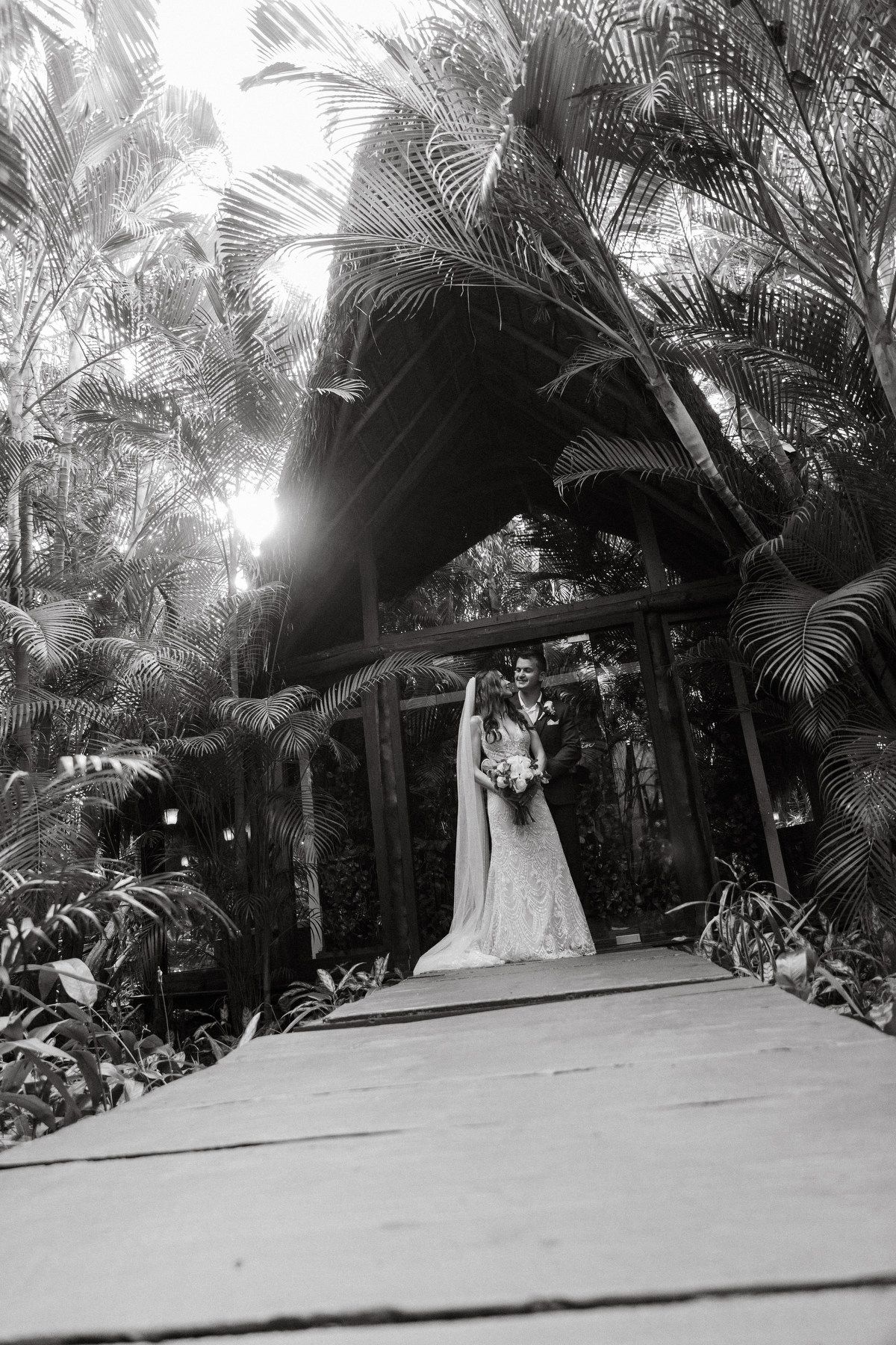 Bride and groom pose under a wooden arch surrounded by lush tropical palm trees in a black and white wedding photo.