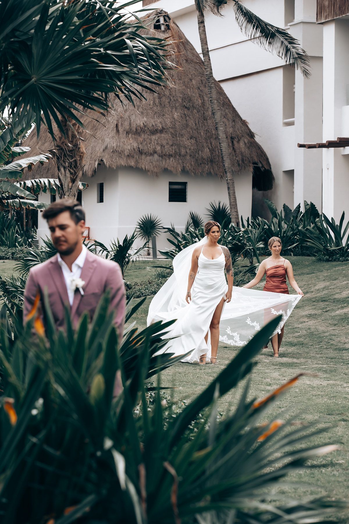 Wedding guests gather on a tropical resort lawn with palm trees and thatched-roof buildings in the background.