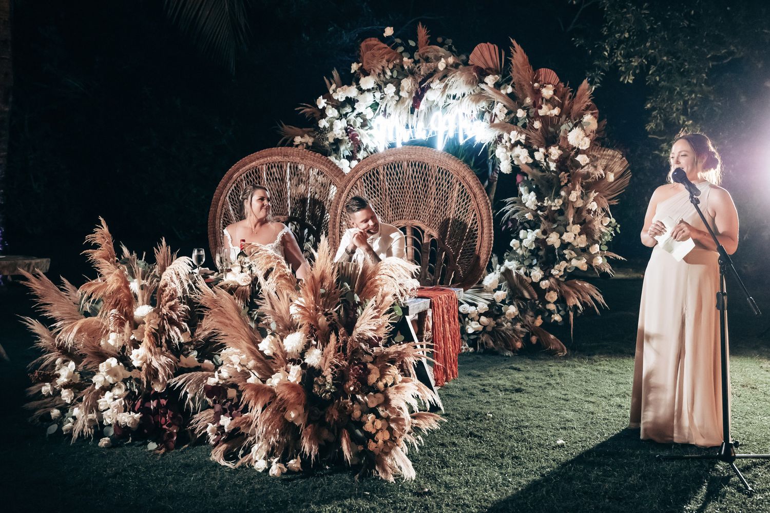 Elegant outdoor wedding ceremony with floral arch, pampas grass arrangements, and bride in white gown at night.