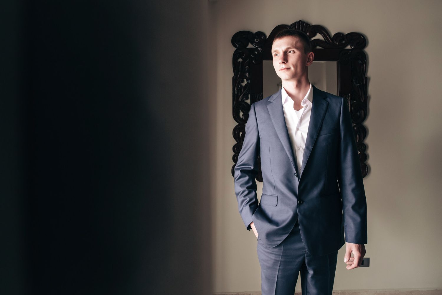 Elegant groom in a navy blue suit standing confidently near an ornate dark mirror frame.