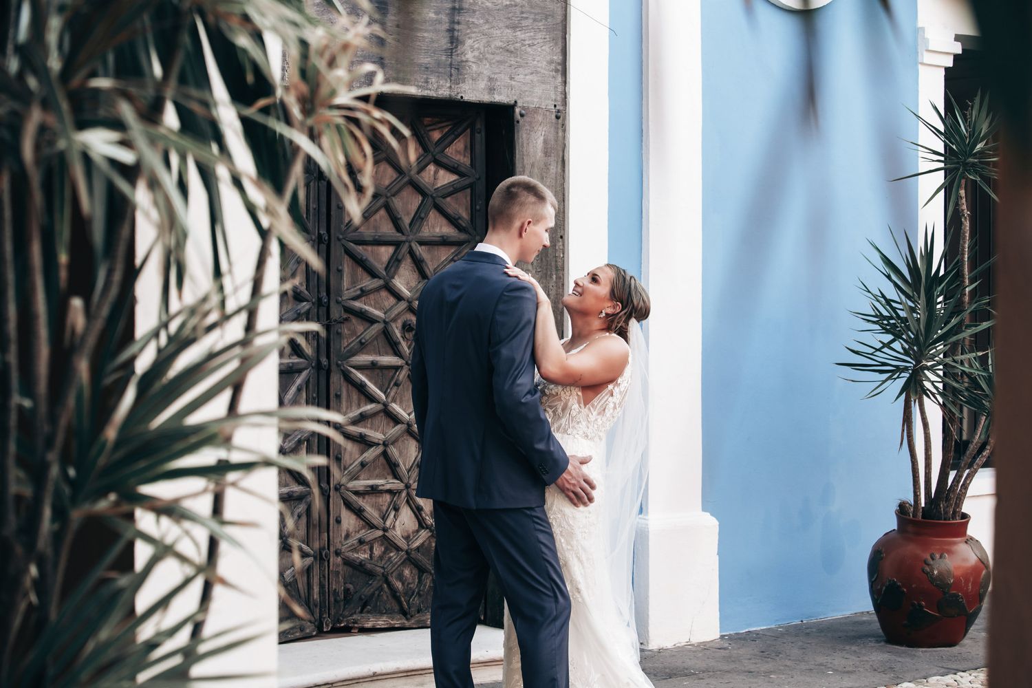 Bride and groom sharing a romantic embrace near a blue wall and ornate wooden door.