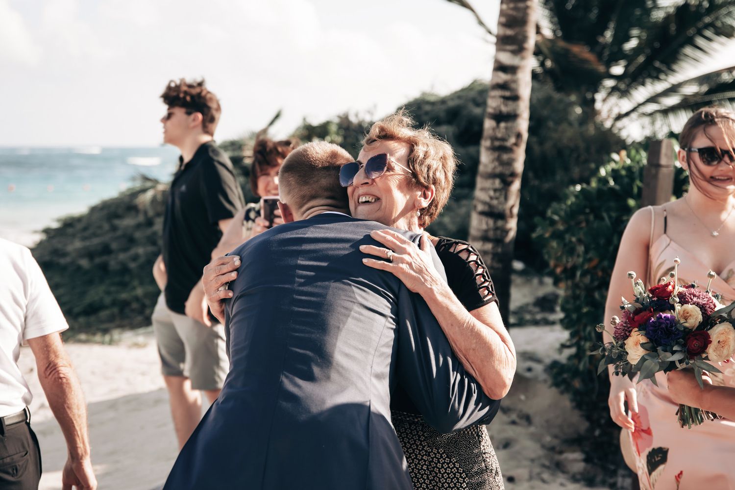 Wedding guest warmly hugs groom near palm trees and rocky beach after outdoor ceremony.