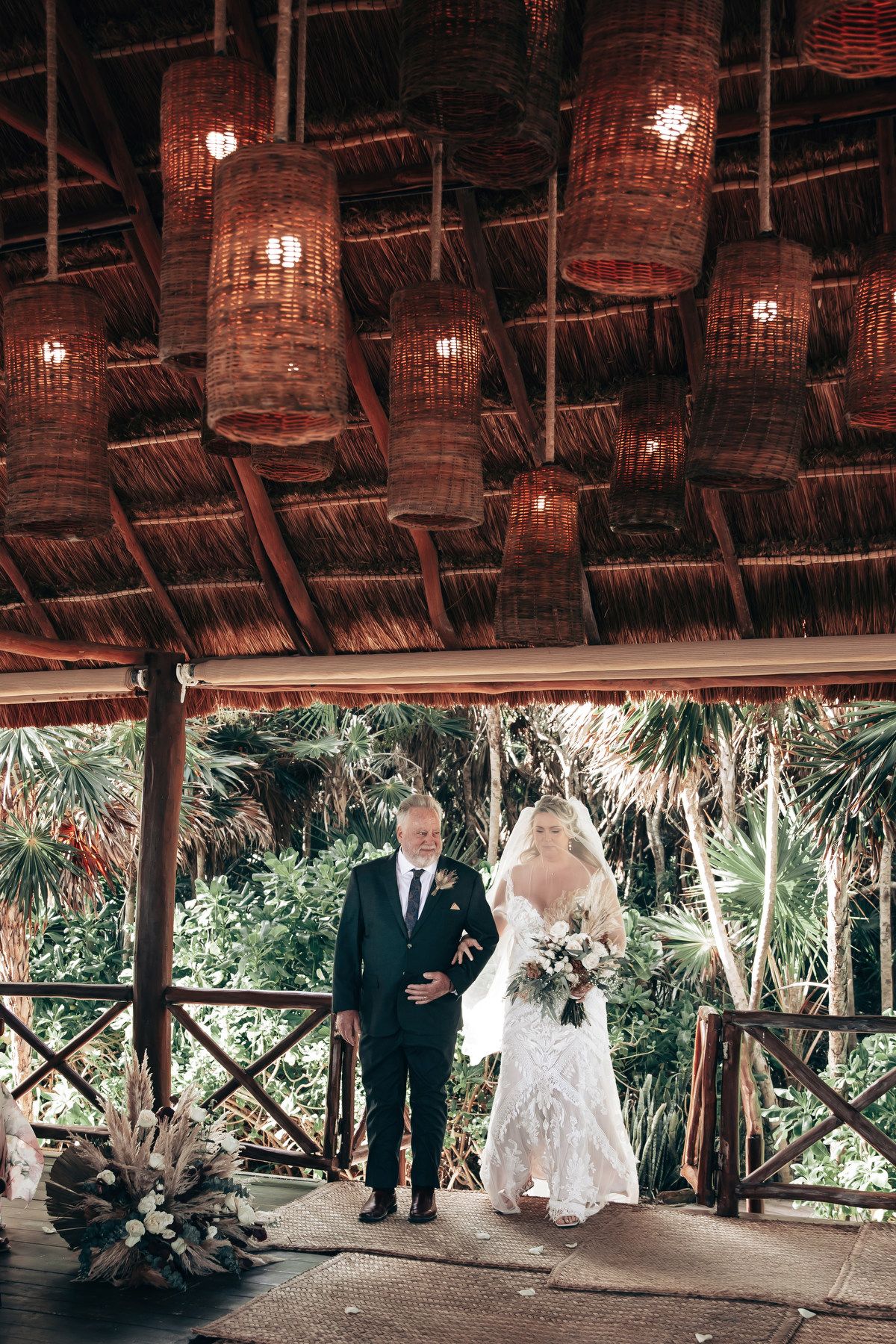 Bride in white lace gown walks with escort under wicker lanterns at tropical outdoor wedding venue at Dreams Tulum