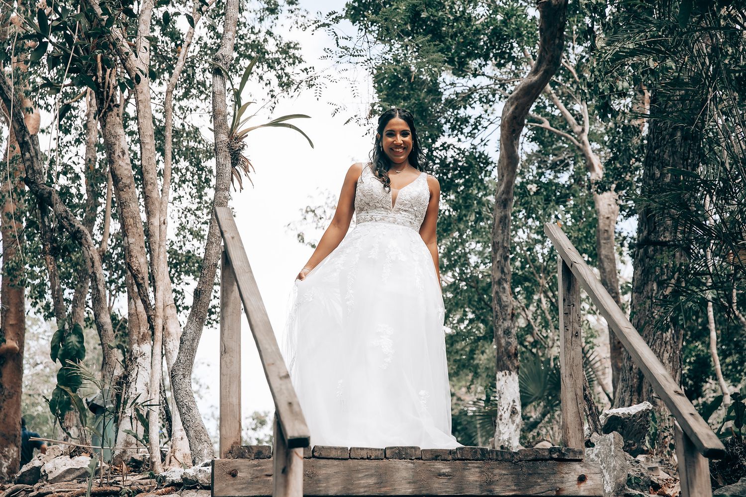 A bride in a flowing white wedding dress poses on wooden steps in a serene forest setting.