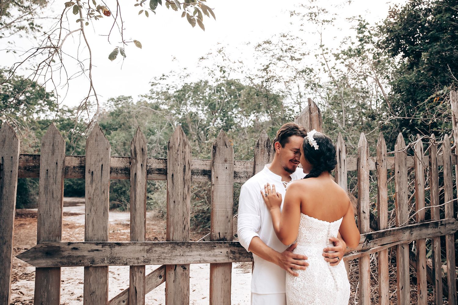 Romantic wedding couple embracing against rustic wooden fence surrounded by lush greenery.