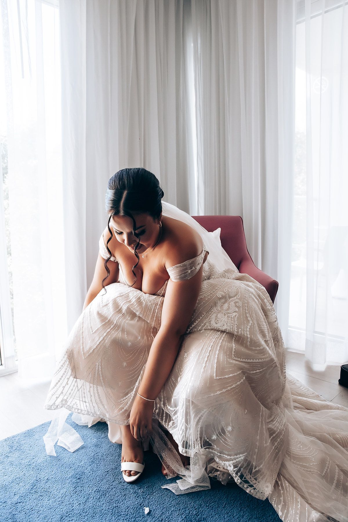 Bride in white wedding gown adjusting her shoe in an elegant room with sheer curtains.