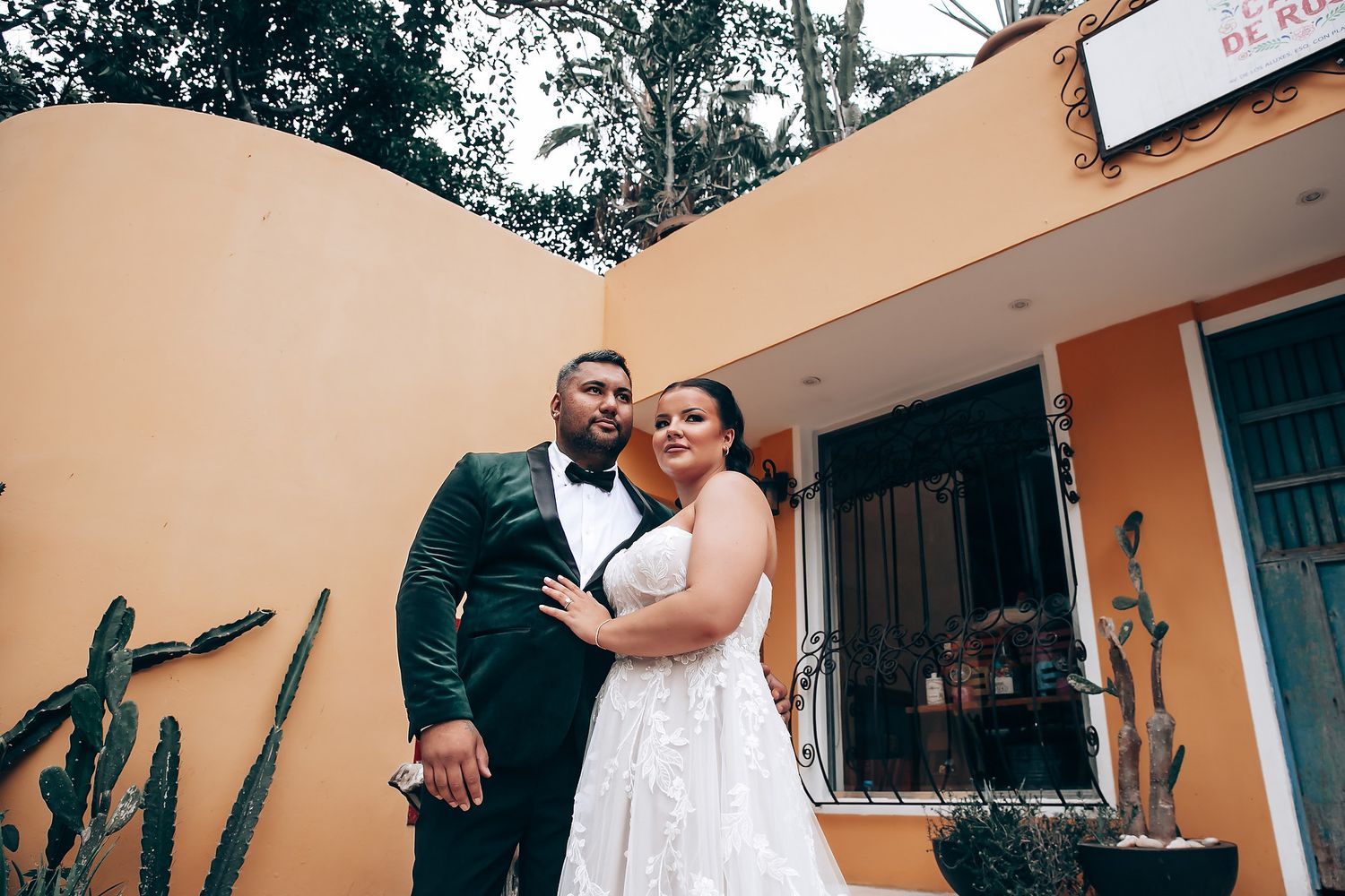 Newlyweds embrace in front of a peach-colored building with tropical plants.