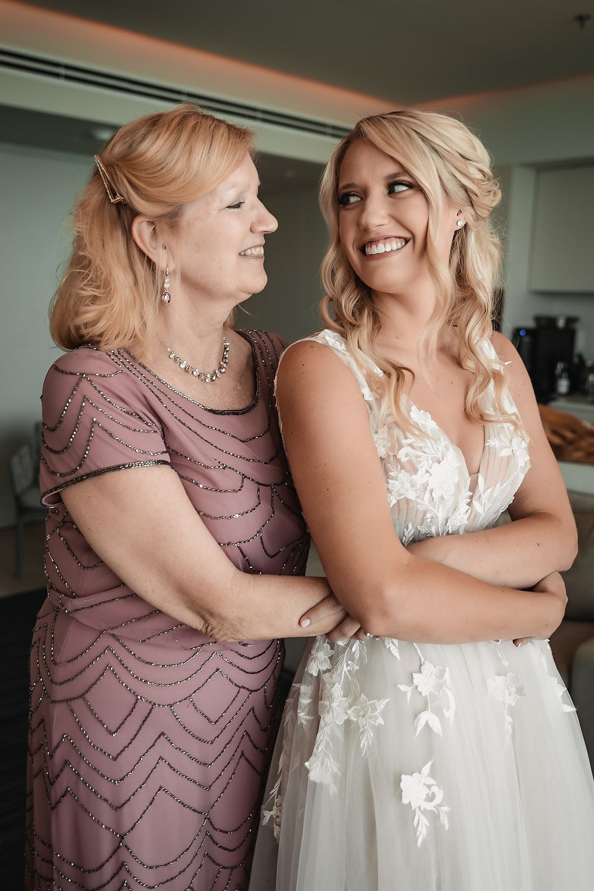 Two women share an emotional moment in matching mauve and white dresses during a wedding preparation.
