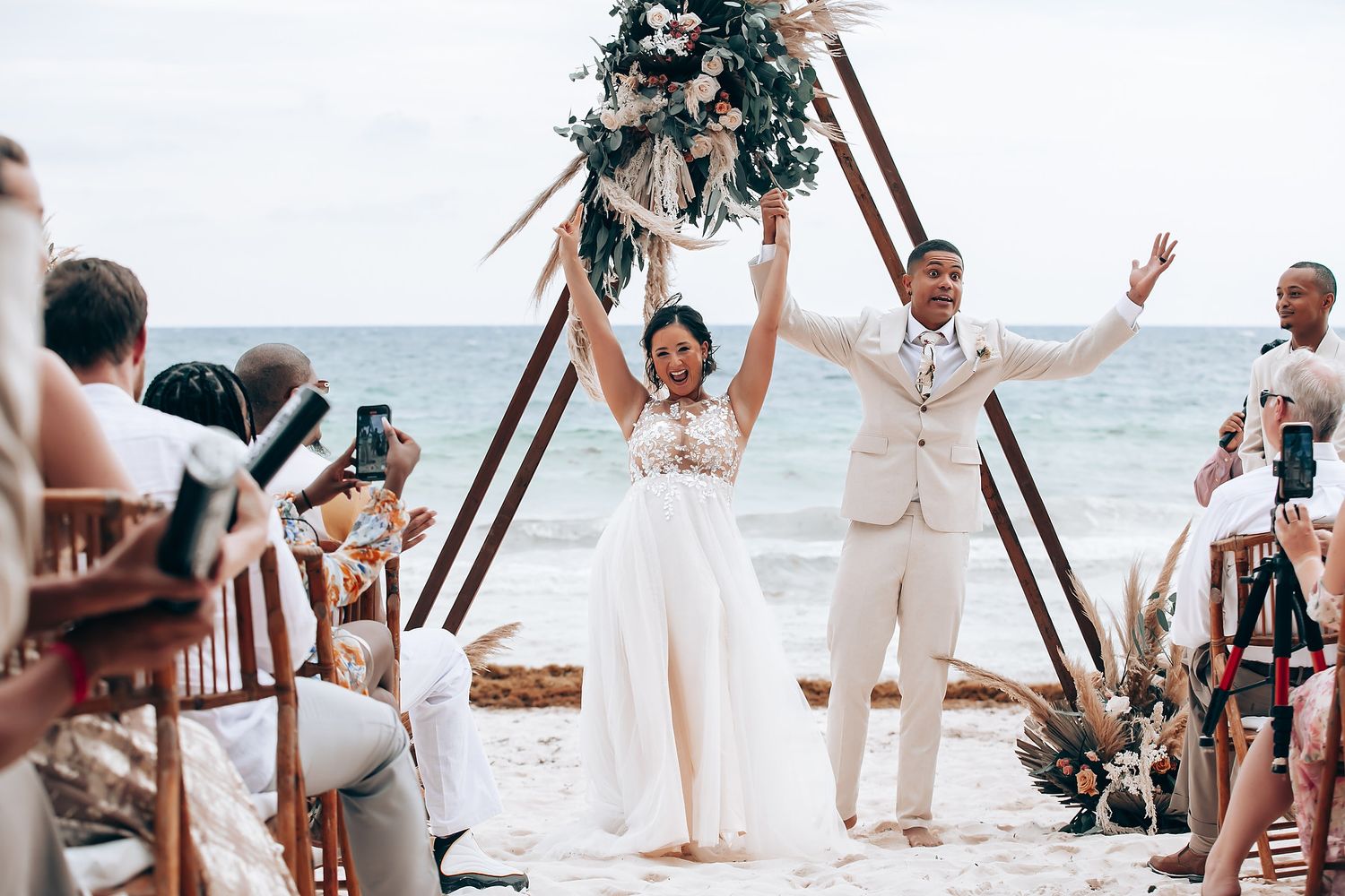 Wedding ceremony celebration on tropical beach with wooden arch and standing guests.