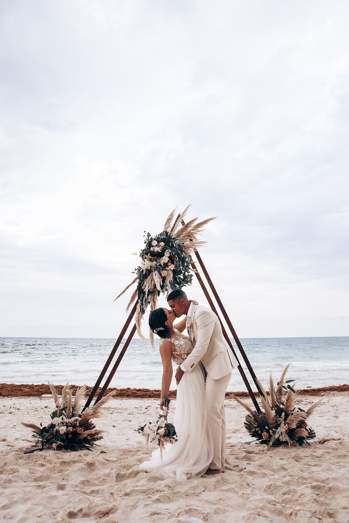 Romantic beach wedding ceremony setup with wooden triangle arch decorated with dried palms and flowers.